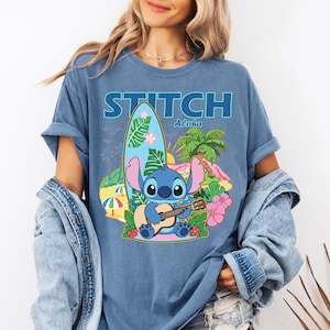 May include: Blue t-shirt featuring a graphic of Stitch playing a ukulele on a tropical beach scene. The word "STITCH" is printed above the image in blue block letters, with "Aloha" below. A surfboard, palm trees, and flowers complete the design.