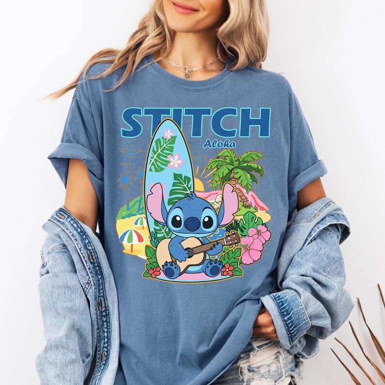 May include: Blue t-shirt featuring a graphic of Stitch playing a ukulele on a tropical beach scene. The word "STITCH" is printed above the image in blue block letters, with "Aloha" below. A surfboard, palm trees, and flowers complete the design.