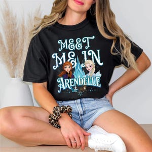 May include: Black t-shirt with the text "MEET ME IN ARENDELLE" in white and teal lettering. The graphic features cartoon illustrations of two princesses and a blue ice crystal. A fun, casual tee.