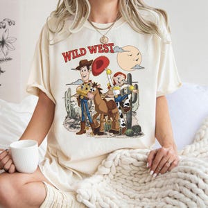 Pu&ograve; includere: T-shirt color crema con grafica Wild West. Il design include personaggi dei cartoni animati, un cavallo, cactus e la scritta "WILD WEST" in rosso. La maglietta ha maniche corte e una vestibilit&agrave; ampia.