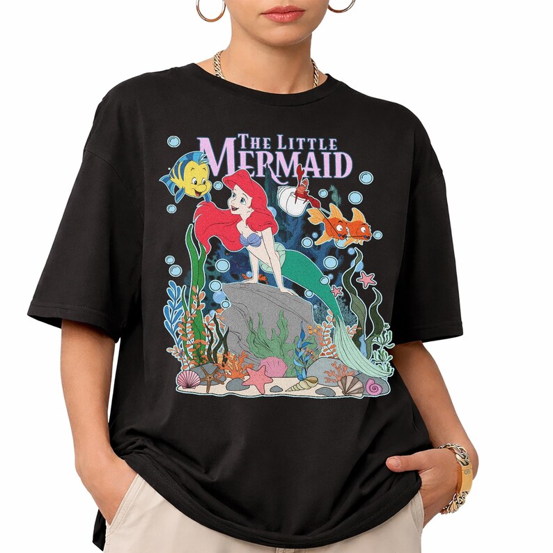 May include: Black t-shirt featuring a colorful graphic of Ariel from The Little Mermaid. The design includes Ariel, Flounder, Sebastian, and other sea creatures, with the words "The Little Mermaid" above the image.