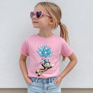 May include: Pink t-shirt featuring a cartoon character holding blue balloons. The character is dressed in a top hat and suit. The shirt has rolled-up sleeves and is worn by a person wearing heart-shaped sunglasses.
