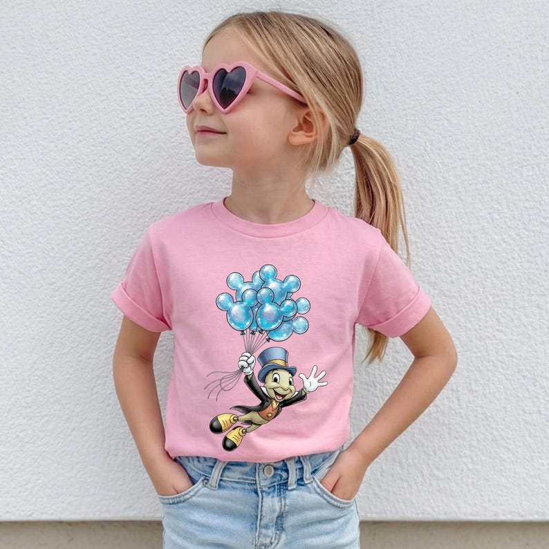 May include: Pink t-shirt featuring a cartoon character holding blue balloons. The character is dressed in a top hat and suit. The shirt has rolled-up sleeves and is worn by a person wearing heart-shaped sunglasses.