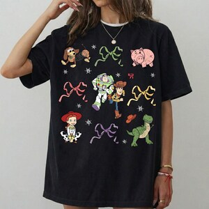 Op de afbeelding: Zwart T-shirt met kleurrijke cartoonfiguren en strikken. Het ontwerp bevat een hond, een varken, Buzz Lightyear, Woody, Jessie en een dinosaurus. Het shirt heeft een ronde hals en korte mouwen.