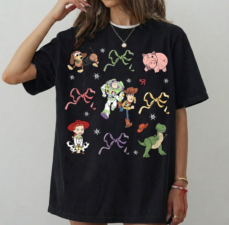 Op de afbeelding: Zwart T-shirt met kleurrijke cartoonfiguren en strikken. Het ontwerp bevat een hond, een varken, Buzz Lightyear, Woody, Jessie en een dinosaurus. Het shirt heeft een ronde hals en korte mouwen.