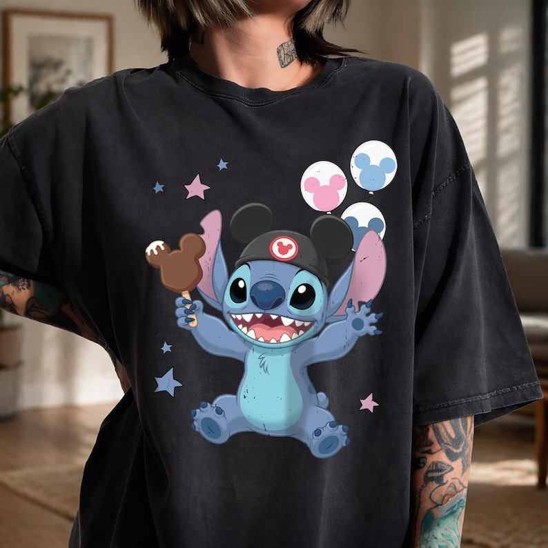 May include: A black short-sleeve t-shirt featuring a graphic of the blue character Stitch wearing a black hat with mouse ears. Stitch holds a brown mouse-shaped ice cream bar, surrounded by pink and blue balloons and small star shapes.