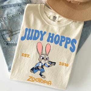 May include: A cream-colored short-sleeve t-shirt featuring a graphic of the character Judy Hopps from Zootopia. The text reads JUDY HOPPS in blue, EST. 2016 in gold, and ZOOTOPIA at the bottom. The shirt has a crew neckline and a Comfort Colors label.