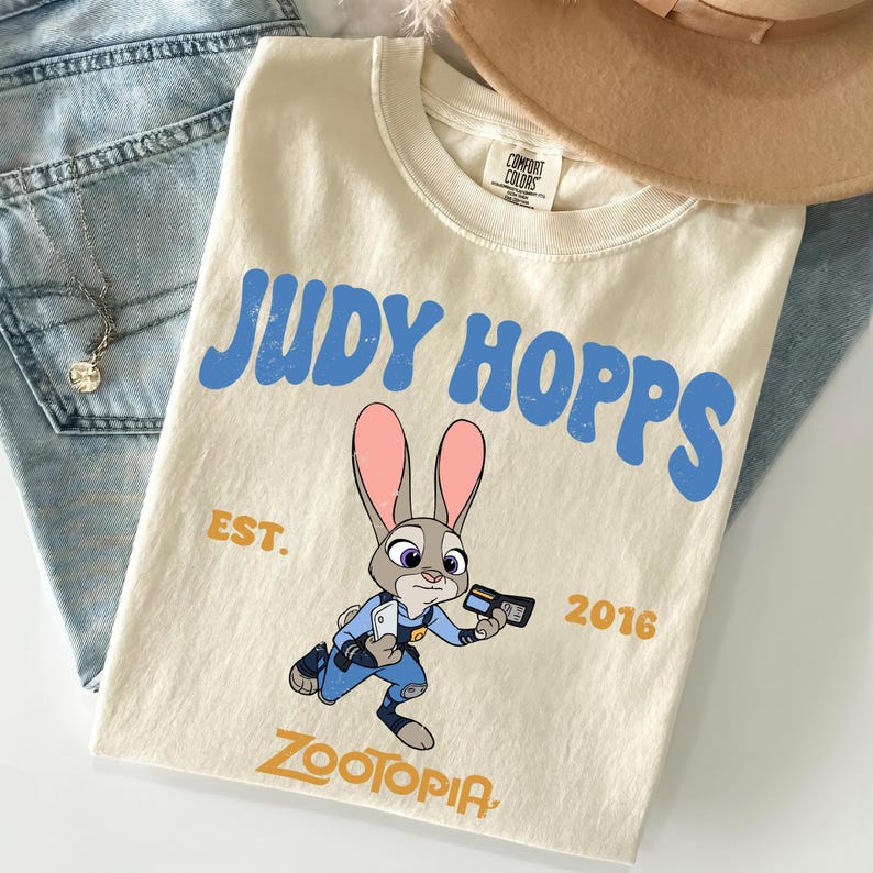 May include: A cream-colored short-sleeve t-shirt featuring a graphic of the character Judy Hopps from Zootopia. The text reads JUDY HOPPS in blue, EST. 2016 in gold, and ZOOTOPIA at the bottom. The shirt has a crew neckline and a Comfort Colors label.