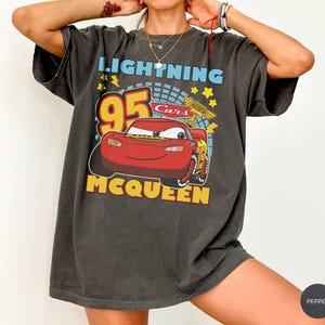 May include: Dark gray t-shirt featuring a graphic of Lightning McQueen from the Cars movie. The design includes the number 95, the words "Lightning McQueen" and "Cars" in yellow and blue. The shirt has short sleeves.