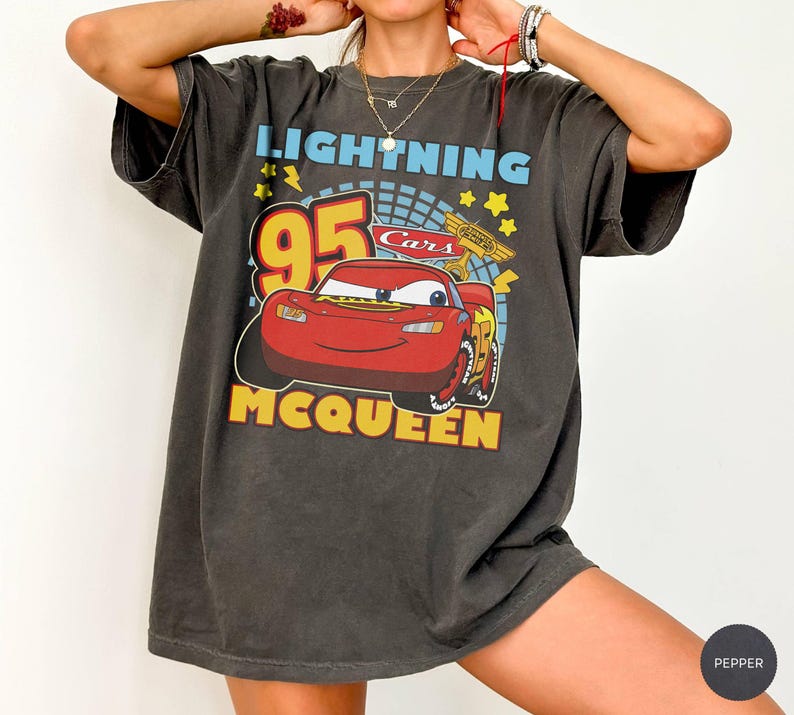 May include: Dark gray t-shirt featuring a graphic of Lightning McQueen from the Cars movie. The design includes the number 95, the words "Lightning McQueen" and "Cars" in yellow and blue. The shirt has short sleeves.