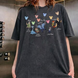 May include: A charcoal grey short-sleeve t-shirt featuring a graphic print of various animated animal characters holding colourful balloons shaped like mouse ears. The text includes names such as Flash, Chief Bogo, Nick Wilde, Gary De Snake, Nibbles Maplestick, and Judy Hopps.