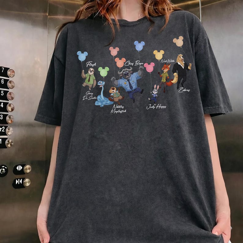 May include: A charcoal grey short-sleeve t-shirt featuring a graphic print of various animated animal characters holding colourful balloons shaped like mouse ears. The text includes names such as Flash, Chief Bogo, Nick Wilde, Gary De Snake, Nibbles Maplestick, and Judy Hopps.