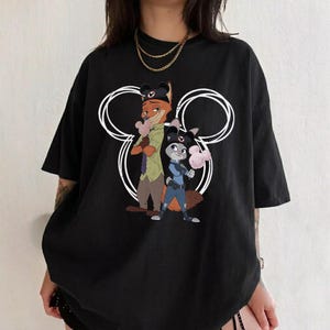 May include: A black short-sleeved t-shirt featuring a graphic print of two animated fox and rabbit characters wearing dark hats. The characters are positioned in front of a large white circular outline resembling mouse ears, each holding a pink treat.