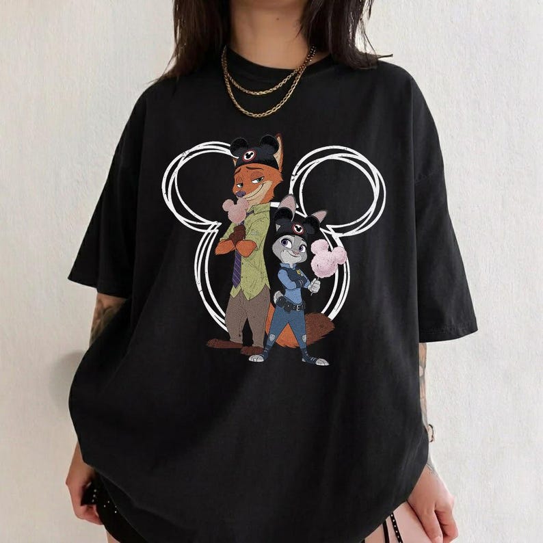 May include: A black short-sleeved t-shirt featuring a graphic print of two animated fox and rabbit characters wearing dark hats. The characters are positioned in front of a large white circular outline resembling mouse ears, each holding a pink treat.
