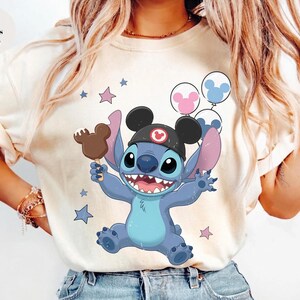 May include: An ivory t-shirt featuring a graphic of the blue character Stitch wearing black mouse ears, holding a brown chocolate-covered treat, and surrounded by pink and blue balloons and stars. The design includes a small red circular icon.