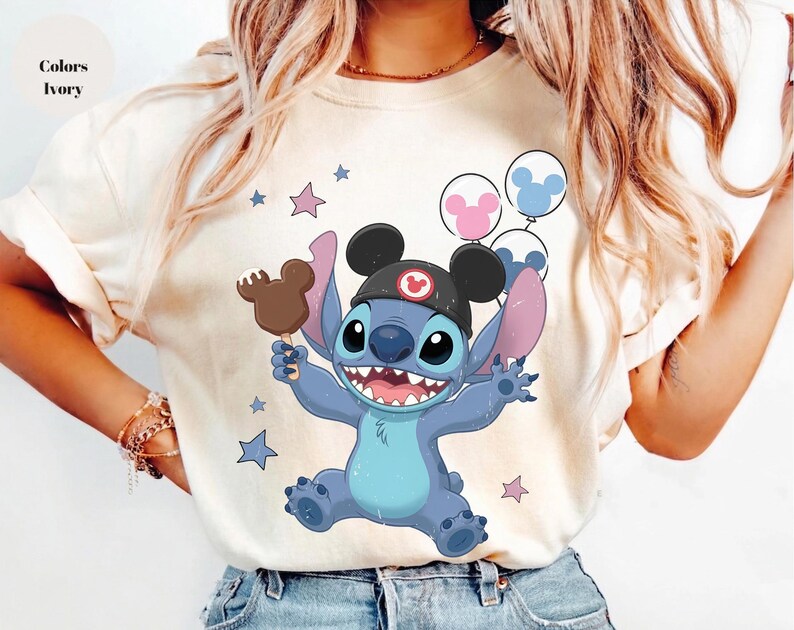 May include: An ivory t-shirt featuring a graphic of the blue character Stitch wearing black mouse ears, holding a brown chocolate-covered treat, and surrounded by pink and blue balloons and stars. The design includes a small red circular icon.