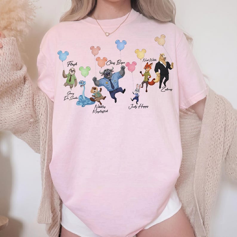 May include: A light pink t-shirt featuring a graphic print of various animated characters, including Flash, Chief Bogo, Nick Wilde, and Judy Hopps, each holding a colourful Mickey Mouse-shaped balloon. The text labels identify each character.