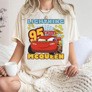May include: Cream-colored t-shirt featuring a graphic of Lightning McQueen from the Cars movie. The design includes the text "LIGHTNING", "95", "McQUEEN", and the Cars logo. The shirt has short sleeves and a relaxed fit.
