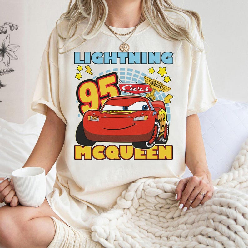 May include: Cream-colored t-shirt featuring a graphic of Lightning McQueen from the Cars movie. The design includes the text "LIGHTNING", "95", "McQUEEN", and the Cars logo. The shirt has short sleeves and a relaxed fit.