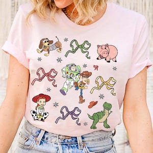 Op de afbeelding: Lichtroze t-shirt met stripfiguren en strikken. Het ontwerp bevat een teckel, een varken, Buzz Lightyear, Woody, Jessie en een groene dinosaurus. Het shirt heeft ook ster- en strik-afbeeldingen.