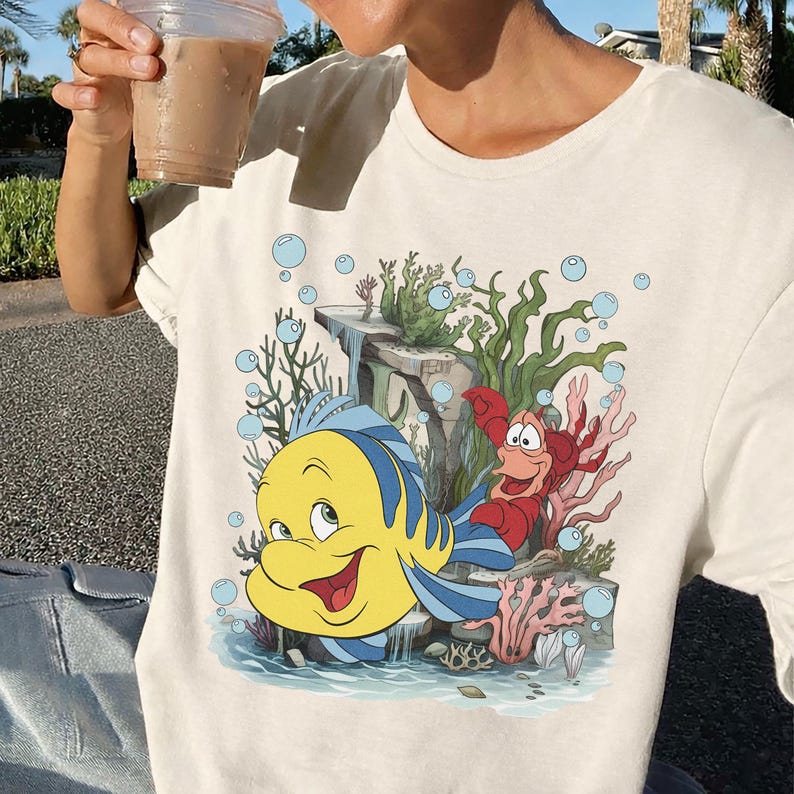 Puede incluir: Camiseta color crema con un gr&aacute;fico colorido de Flounder y Sebasti&aacute;n de La Sirenita. El dise&ntilde;o incluye burbujas azules, coral y plantas acu&aacute;ticas. Los personajes est&aacute;n ambientados frente a una cascada.