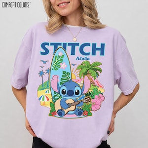 May include: A light purple t-shirt with a graphic of Stitch playing a ukulele in front of a surfboard, palm trees, and flowers. The word "STITCH" is above the graphic in blue letters, with "Aloha" below.