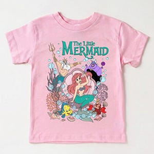 Puede incluir: Camiseta rosa con un gr&aacute;fico de Ariel de La Sirenita. El dise&ntilde;o incluye a Ariel, el Rey Trit&oacute;n, &Uacute;rsula, Flounder y Sebasti&aacute;n, con las palabras "The Little Mermaid" en verde azulado. La camiseta es de material suave.