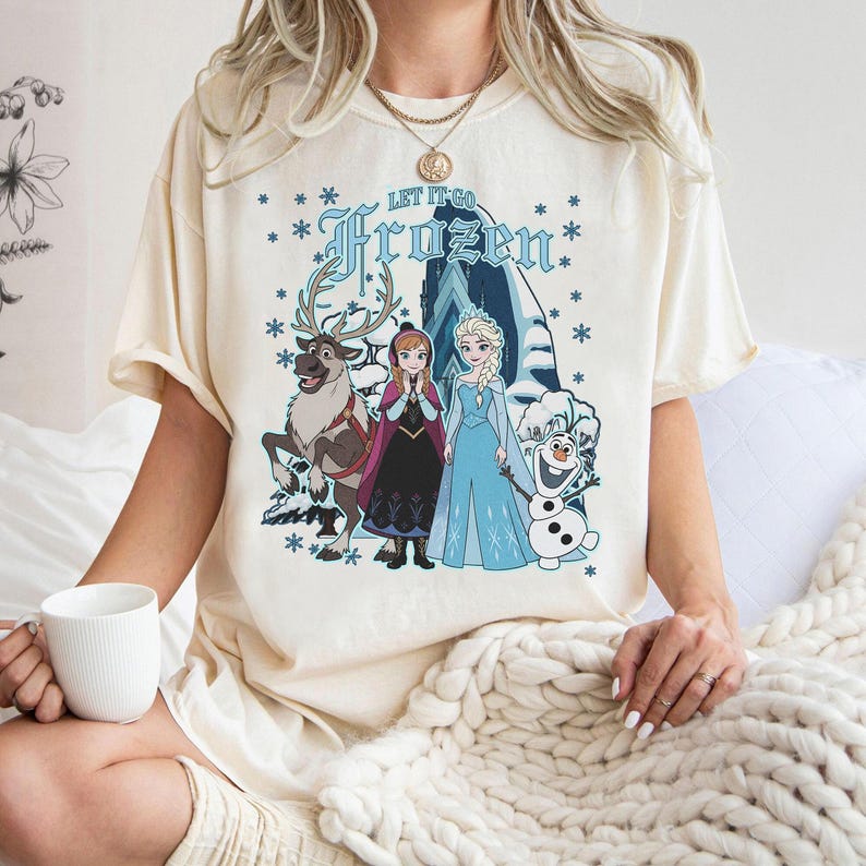 May include: Cream-colored t-shirt featuring the text "Let It Go Frozen" in blue, with illustrations of characters from the movie Frozen, including Anna, Elsa, Olaf, and Sven. The design is surrounded by blue snowflakes.