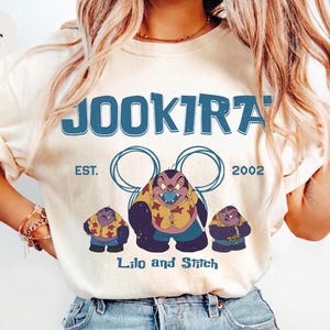 Puede incluir: Una camiseta color marfil con un estampado gr&aacute;fico azul con el texto JOOKIRA, EST. 2002 y Lilo and Stitch. El dise&ntilde;o incluye tres personajes de dibujos animados con camisas estampadas en amarillo y rojo, bajo dos contornos circulares azules.