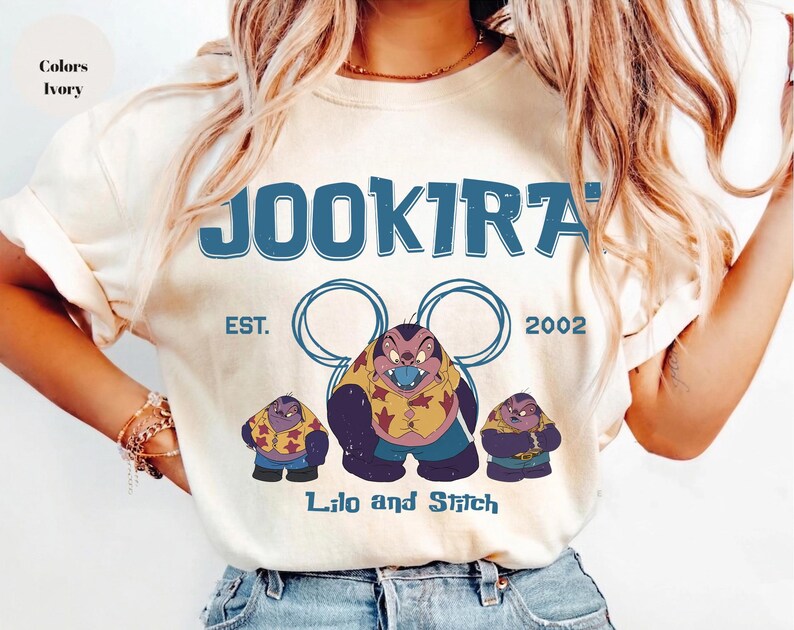 Puede incluir: Una camiseta color marfil con un estampado gr&aacute;fico azul con el texto JOOKIRA, EST. 2002 y Lilo and Stitch. El dise&ntilde;o incluye tres personajes de dibujos animados con camisas estampadas en amarillo y rojo, bajo dos contornos circulares azules.