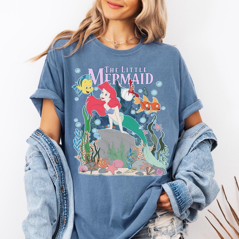 May include: Blue t-shirt featuring a graphic of Ariel from The Little Mermaid. The design includes Ariel, Flounder, and other sea creatures. The words "The Little Mermaid" are printed across the top in pink and white.