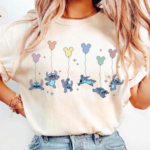 May include: An ivory short-sleeved t-shirt featuring a graphic print of the blue character Stitch from Lilo and Stitch. The design shows multiple versions of the character holding onto colorful balloons shaped like mouse ears with small stars.