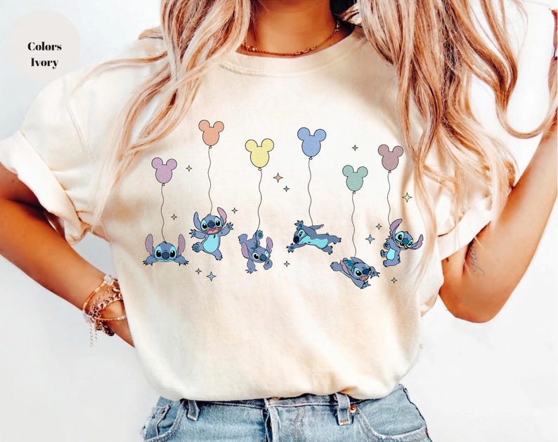 May include: An ivory short-sleeved t-shirt featuring a graphic print of the blue character Stitch from Lilo and Stitch. The design shows multiple versions of the character holding onto colorful balloons shaped like mouse ears with small stars.