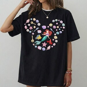 May include: Black t-shirt featuring a colorful design of a mermaid, seashells, and aquatic elements arranged in a Mickey Mouse head shape. The mermaid has red hair and a green tail. The design includes a yellow fish and a red lobster.