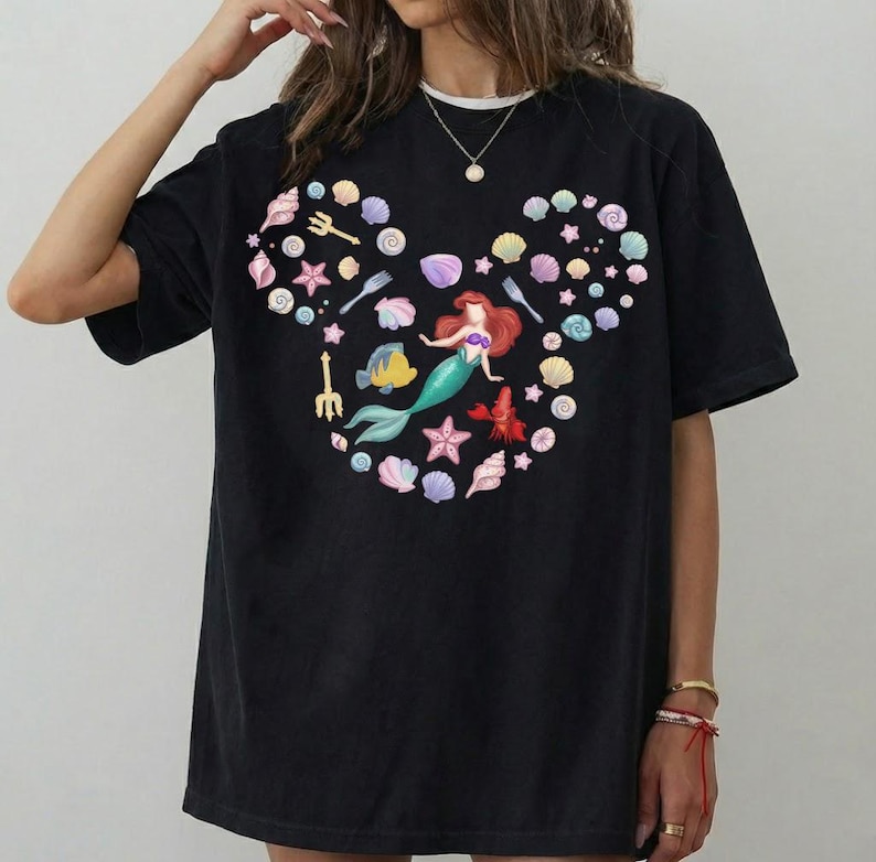 May include: Black t-shirt featuring a colorful design of a mermaid, seashells, and aquatic elements arranged in a Mickey Mouse head shape. The mermaid has red hair and a green tail. The design includes a yellow fish and a red lobster.