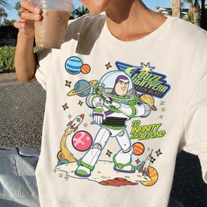 May include: Cream-colored t-shirt featuring a colorful graphic of Buzz Lightyear from Toy Story. The design includes the text "Buzz Lightyear Laser Blast" and "To Infinity and Beyond" with planets, rockets, and stars.