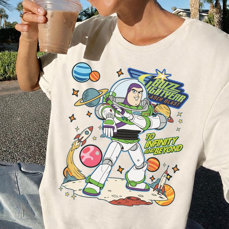 May include: Cream-colored t-shirt featuring a colorful graphic of Buzz Lightyear from Toy Story. The design includes the text "Buzz Lightyear Laser Blast" and "To Infinity and Beyond" with planets, rockets, and stars.