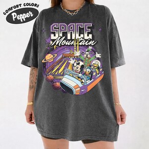 May include: Dark grey Comfort Colors t-shirt with a colourful graphic of Mickey Mouse, Donald Duck, Goofy, and Pluto on the Space Mountain ride. The shirt has the words "SPACE Mountain" in a purple and white design.