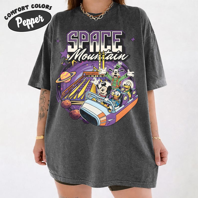 May include: Dark grey Comfort Colors t-shirt with a colourful graphic of Mickey Mouse, Donald Duck, Goofy, and Pluto on the Space Mountain ride. The shirt has the words "SPACE Mountain" in a purple and white design.