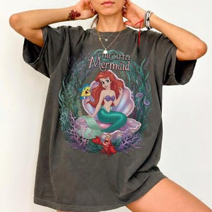 May include: Dark gray t-shirt featuring a graphic of Ariel from The Little Mermaid. Ariel is depicted with red hair, a purple seashell bra, and a green tail, seated in a seashell. The shirt includes the text "The Little Mermaid".