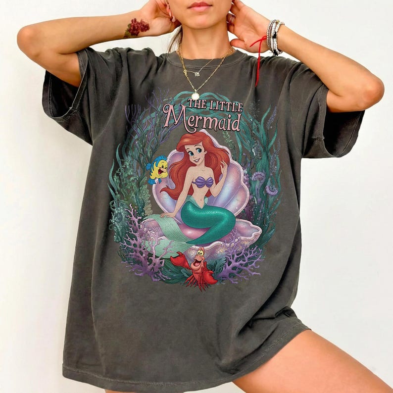 May include: Dark gray t-shirt featuring a graphic of Ariel from The Little Mermaid. Ariel is depicted with red hair, a purple seashell bra, and a green tail, seated in a seashell. The shirt includes the text "The Little Mermaid".