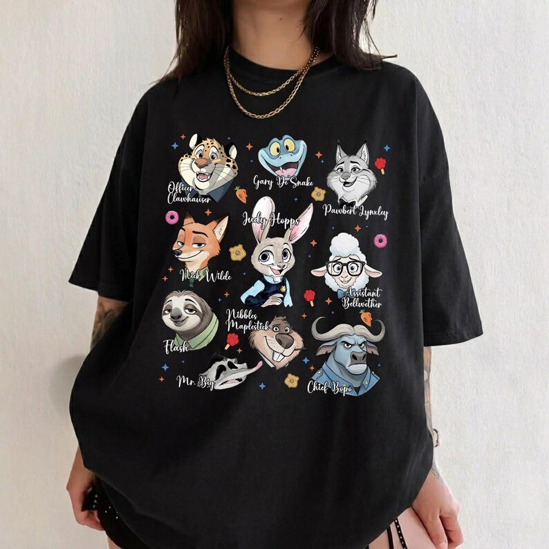 May include: A black short-sleeved t-shirt featuring a graphic print of various animated animal characters from Zootopia, including Judy Hopps, Nick Wilde, Flash, and Chief Bogo, with their names printed in white script text alongside small colorful icons.