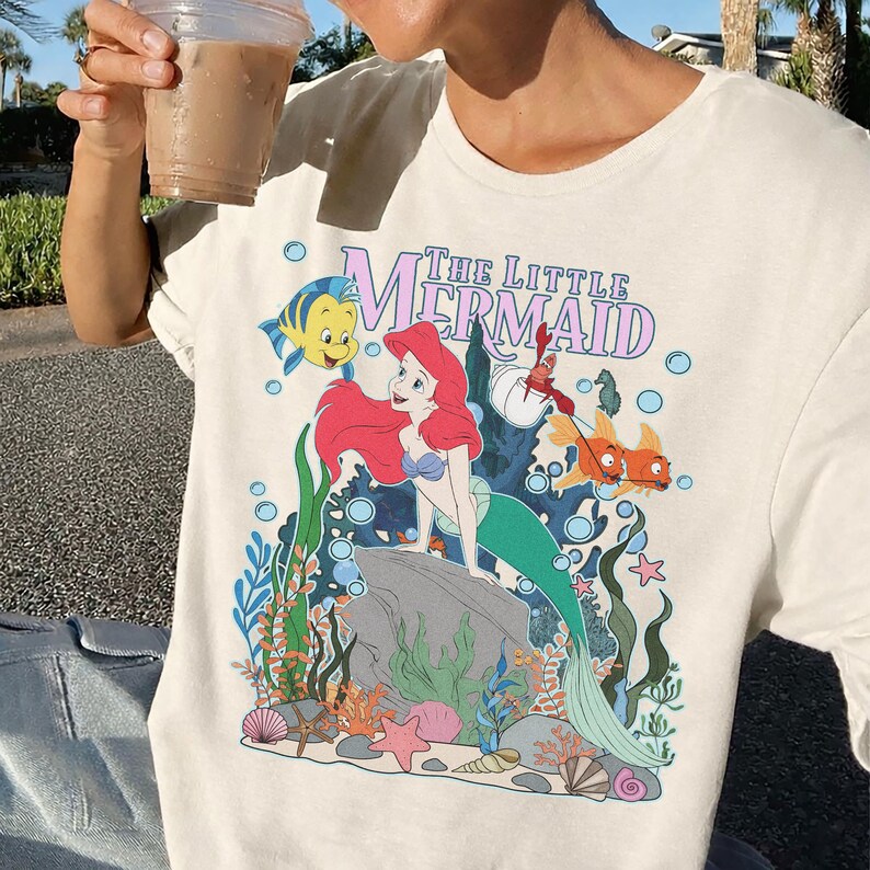 May include: Cream-colored t-shirt featuring a colorful graphic of Ariel from The Little Mermaid. The design includes the text "The Little Mermaid" in a purple font, along with illustrations of Flounder, Sebastian, and other sea creatures.