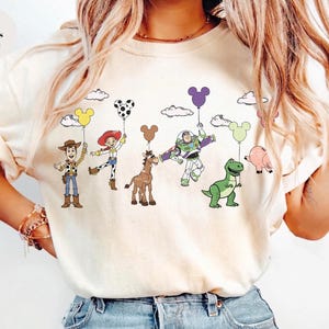 Op de afbeelding: Ivoor kleurig T-shirt met een kleurrijke afbeelding van stripfiguren die ballonvormige ballonnen vasthouden. De figuren zijn Woody, Jessie, Buzz Lightyear, een paard, een dinosaurus en een varken. De tekst "Colors Ivory" staat linksboven.