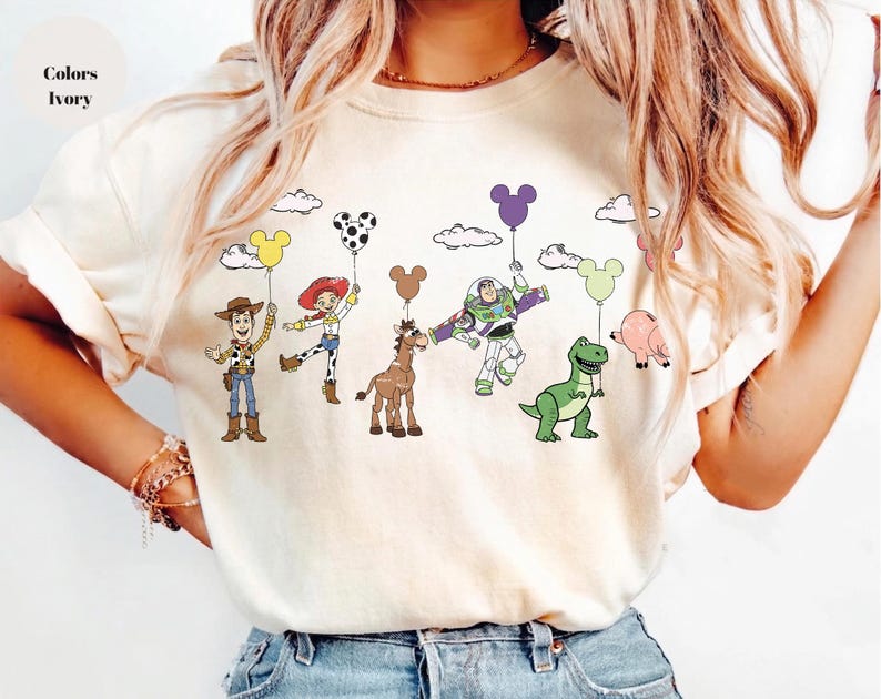 Op de afbeelding: Ivoor kleurig T-shirt met een kleurrijke afbeelding van stripfiguren die ballonvormige ballonnen vasthouden. De figuren zijn Woody, Jessie, Buzz Lightyear, een paard, een dinosaurus en een varken. De tekst "Colors Ivory" staat linksboven.