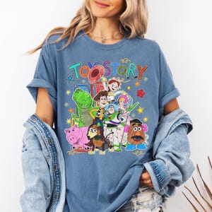Może przedstawiać: Niebieska koszulka z kolorową grafiką postaci z Toy Story, w tym Chudego, Buzz'a Astrala i Rexa. Tekst "TOY STORY" jest napisany nad postaciami zabawną czcionką. Koszulka ma podwinięte rękawy.