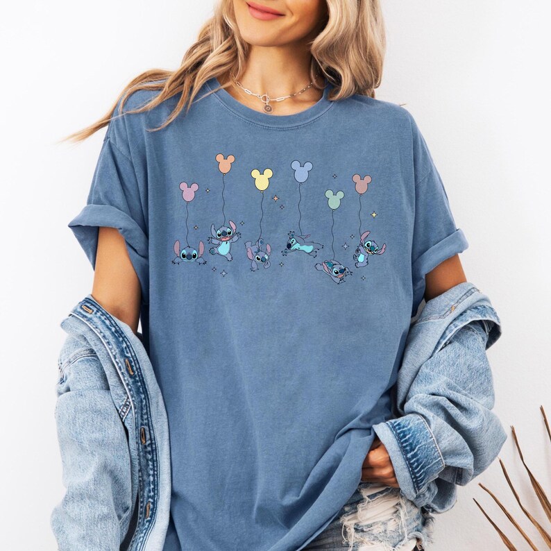 May include: A blue short-sleeve t-shirt featuring a graphic print of the character Stitch in various poses, each holding a pastel-colored balloon shaped like a mouse head, accented with small star illustrations scattered across the chest area.
