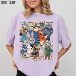Op de afbeelding: Een lichtpaars Comfort Colors T-shirt met een kleurrijke afbeelding van personages uit de film Zootopia, waaronder Judy Hopps en Nick Wilde. Het ontwerp toont verschillende personages uit de film. Het T-shirt heeft korte mouwen.
