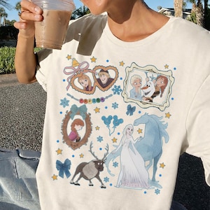 Puede incluir: Camiseta de color crema con un gr&aacute;fico colorido de personajes y elementos de la pel&iacute;cula animada "Frozen". El dise&ntilde;o incluye retratos enmarcados, estrellas, copos de nieve y la palabra "FROZEN".