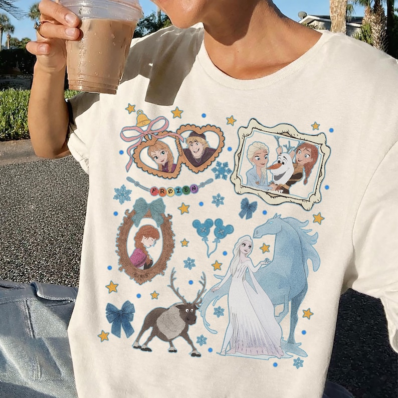 Puede incluir: Camiseta de color crema con un gr&aacute;fico colorido de personajes y elementos de la pel&iacute;cula animada "Frozen". El dise&ntilde;o incluye retratos enmarcados, estrellas, copos de nieve y la palabra "FROZEN".
